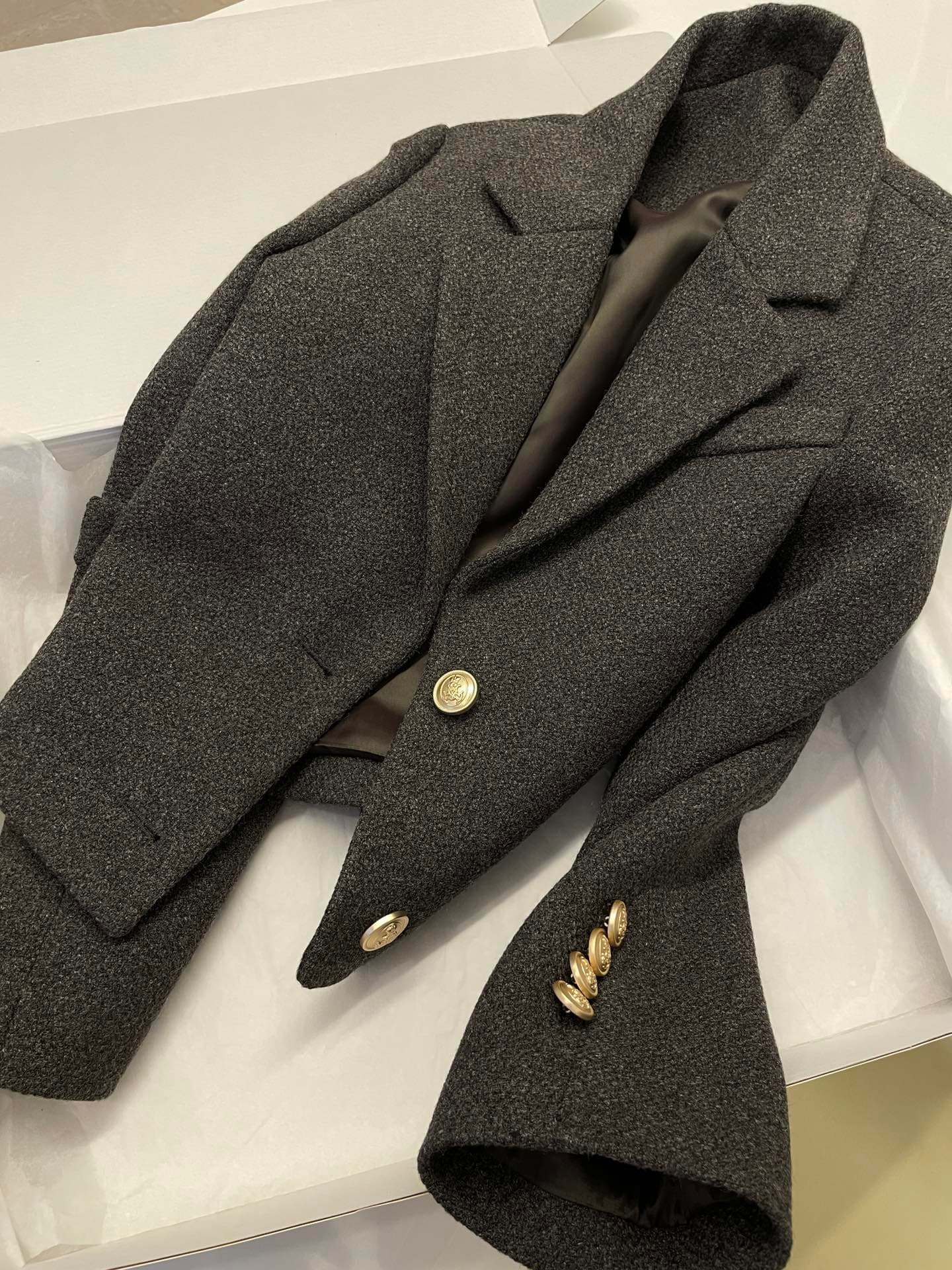 A finished charcoal-grey wool blazer with ornate gold buttons, folded in its packaging box — a brand garment ready to ship